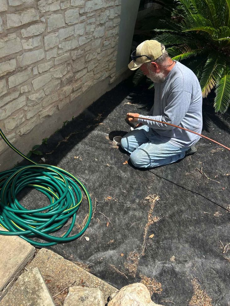 Dave Flynn of Sewer TV Hydro Jetting and Plumbing of San Antonio kneeling beside a stone wall, preparing plumbing equipment next to a coiled green hose on a black ground cover